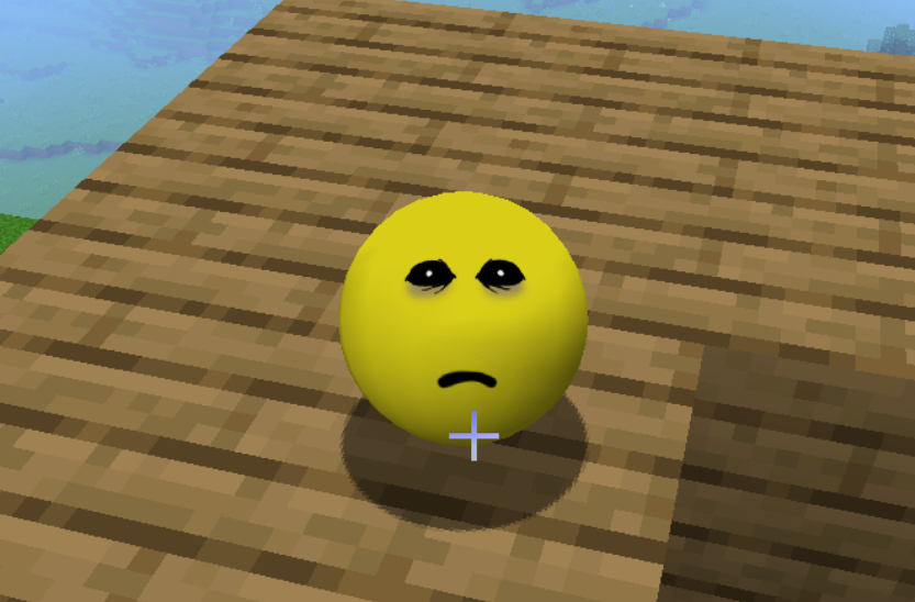 A smooth bright-yellow sphere sits near the center of a first-person view, resting on brown pixelated wooden planks. It has a simple cartoon face: two black oval eyes with small white highlights, angled into a worried or sad shape, and a short black frown. A soft dark shadow sits under the ball. A pale plus-shaped crosshair overlaps the lower edge of the sphere. The planks continue into the distance in a blocky grid. In the upper left there is pixelated blue water, a strip of sky, and a small patch of green. The ball is smoothly shaded, unlike the chunky textures around it.