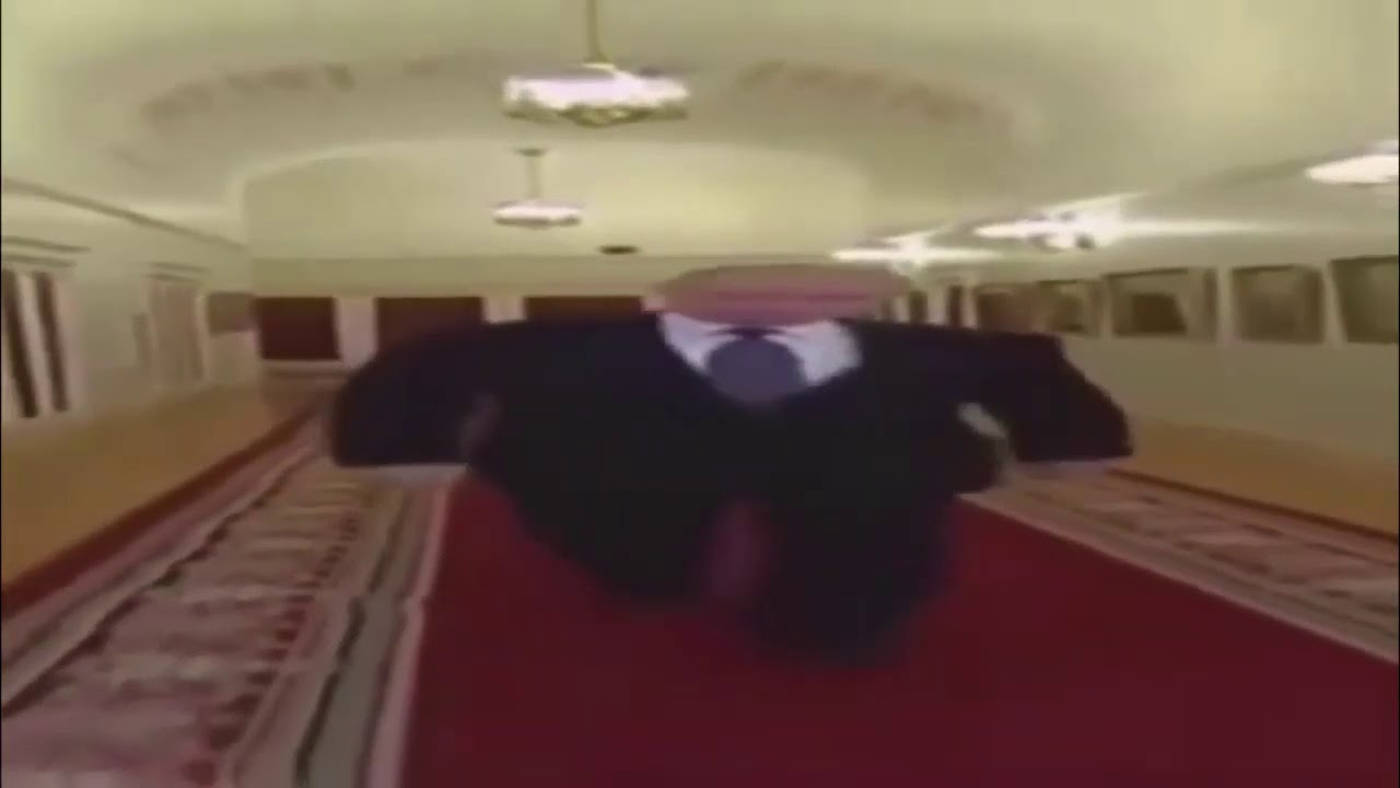 A blurry wide-angle still shows a person in a dark suit, white shirt, and dark tie striding toward the camera with both arms stretched out. The figure looks unnaturally wide. They walk a long red carpet through a pale grand hallway lined with doorways, patterned carpet borders, and ceiling chandeliers. The face is not readable.